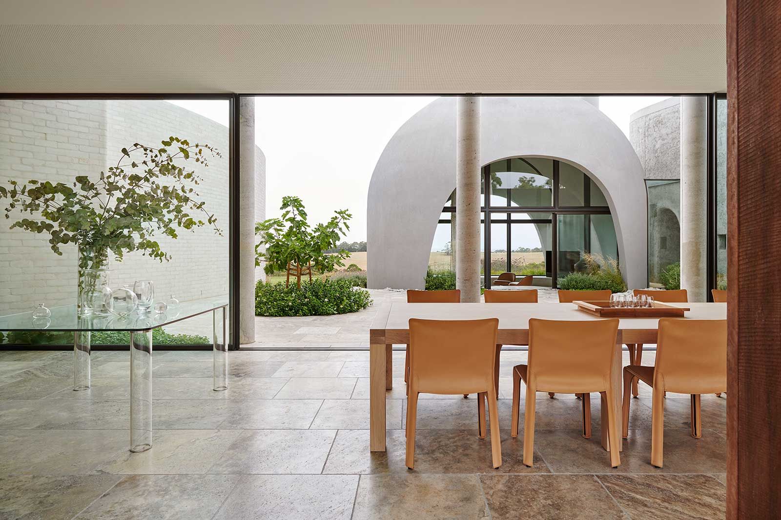 Aus dem Esszimmer schweift der Blick über den Innenhof zum «Round Room» und den sanften Hügeln dahinter. Ein Beistelltisch von Glas Italia ­mit Glasgefässen und Eukalyptusblättern flankiert den massgefertigten Esstisch aus Eiche von Motto Furniture & Cabinetry mit Stühlen von Mario Bellini aus den 1970er-Jahren.